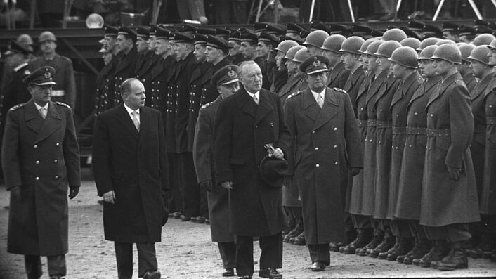 Bundeskanzler Konrad Adenauer und Verteidigungsminister Theodor Blank schreiten bei ihrem Besuch in Andernach zusammen mit drei hochrangigen Bundeswehroffiziere eine angetretene Formation von Soldaten des Heeres und der Marine ab. Im Hintergrund stehen Pressefotografen.