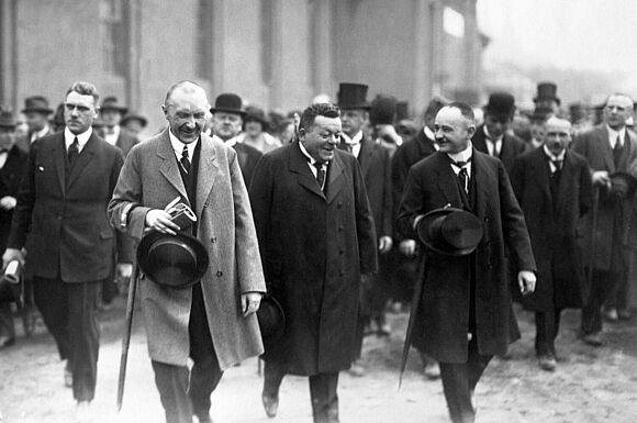 Eröffnung der ersten Kölner Messe 1924 Konrad Adenauer, Friedrich Ebert und Wilhelm Marx laufen lächelnd nebeneinander. Im Hintergrund sind mehrere Menschen in Anzügen und zum Teil mit Hüten zu sehen.