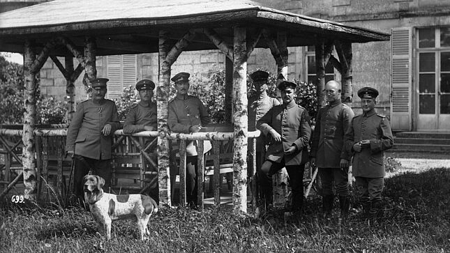 Eine Gruppe von sieben Männern in militärischen Uniformen steht unter einem Unterstand auf einer Wiese und blickt in die Kamera. Im Vordergrund steht ein Hund.