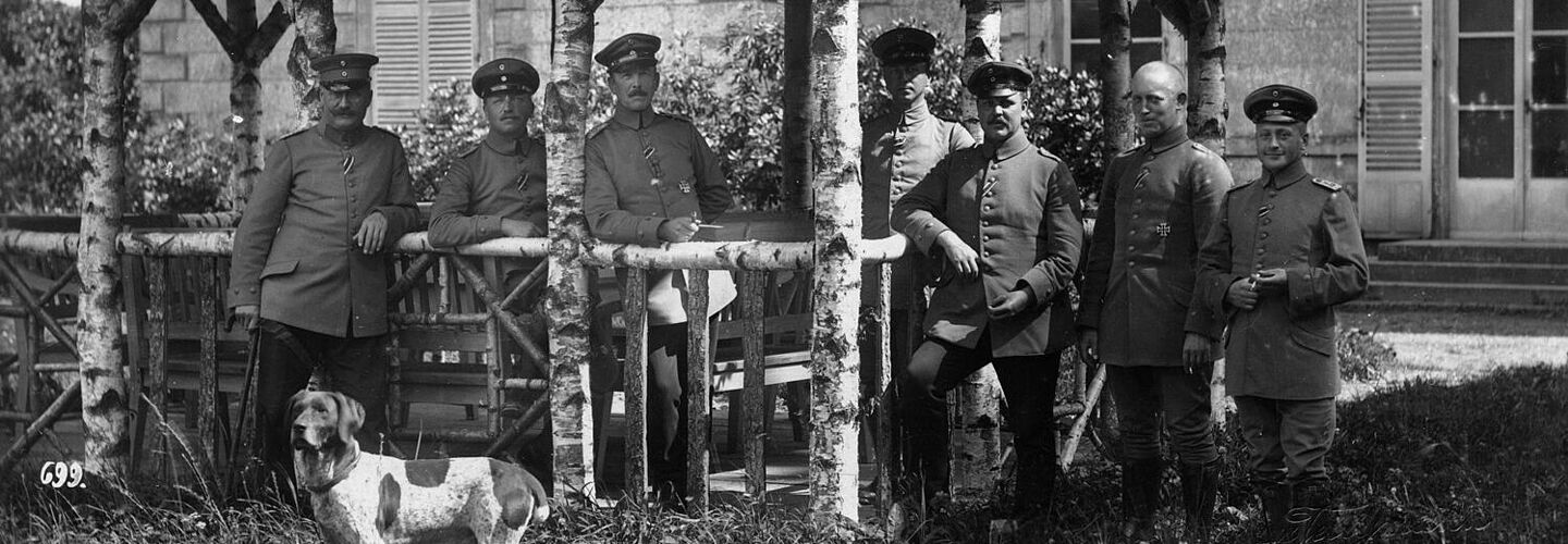 Eine Gruppe von sieben Männern in militärischen Uniformen steht unter einem Unterstand auf einer Wiese und blickt in die Kamera. Im Vordergrund steht ein Hund.