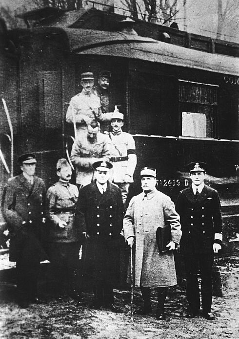 Neun Herren in Uniformen stehen vor und auf einem Eisenbahnwaggon. Ein Mann hält einen Gehstock in der rechten, eine Mappe in der linken Hand. Es handelt sich um Marschall Ferdinand Foch. Das Schwarz-Weiß-Foto wurde im November 1918 in Compiègne, Frankreich, aufgenommen.