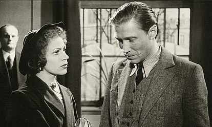 Das Bild zeigt eine Szene aus dem Film 'Affaire Blume' von 1948. Ein Mann und eine Frau stehen halbschräg nebeneinander, der Mann hat den Blick gesenkt, während die Frau zu ihm aufschaut.