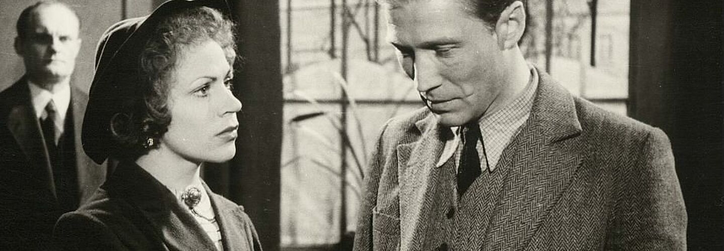 Das Bild zeigt eine Szene aus dem Film 'Affaire Blume' von 1948. Ein Mann und eine Frau stehen halbschräg nebeneinander, der Mann hat den Blick gesenkt, während die Frau zu ihm aufschaut.