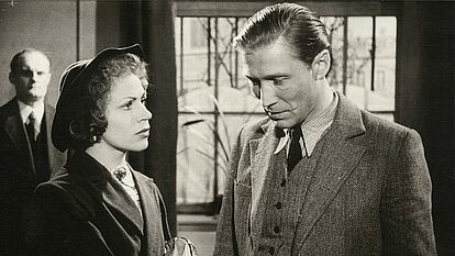 Das Bild zeigt eine Szene aus dem Film 'Affaire Blume' von 1948. Ein Mann und eine Frau stehen halbschräg nebeneinander, der Mann hat den Blick gesenkt, während die Frau zu ihm aufschaut.