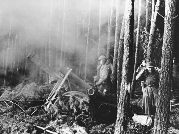 Schweres Infanteriegeschütz im Hürtgenwald Ein schweres Infanteriegeschütz der Wehrmacht steht zwischen Bäumen und feuert. Daneben stehen fünf Angehörige der Wehrmacht, von denen sich einige die Ohren zuhalten.