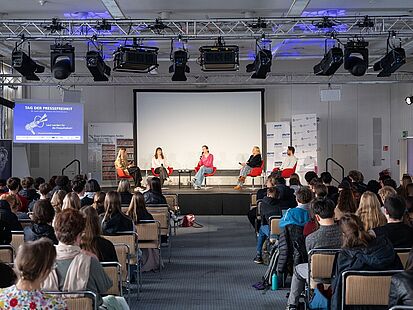 Tag der Pressefreiheit 2026 Auf einem Podium sitzen fünf Personen mit Mikrofonen. Vor dem Podium sitzen Gäste auf Stühlen.
