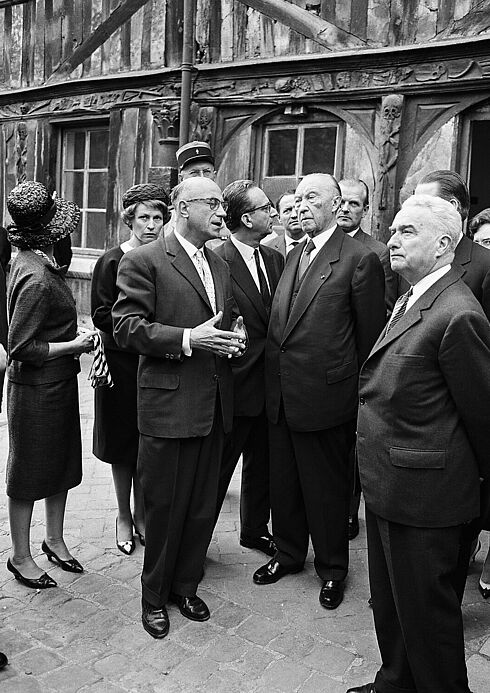 Adenauer sowie viele weitere formell gekleidete Männer und Frauen stehen vor einem alten Gebäude mit Holzverzierungen. Sie hören einem sprechenden Herrn zu, einige schauen sich um.