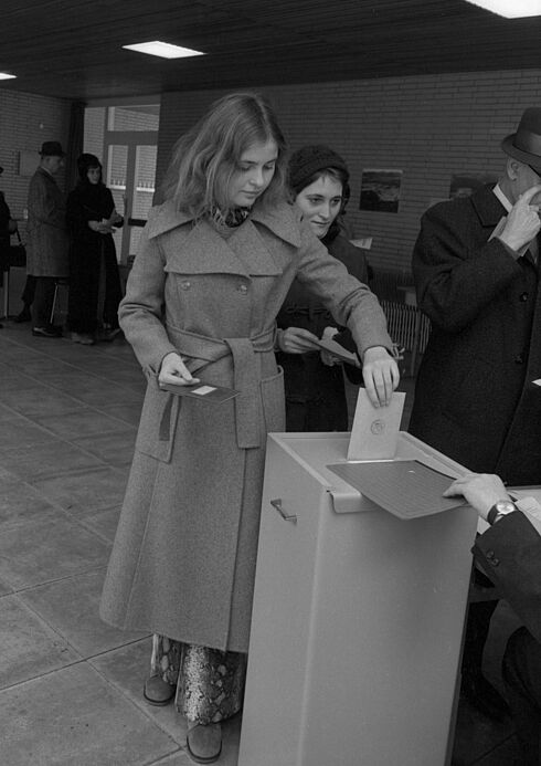 Schwarz-Weiß-Aufnahme in einem Wahllokal, während eine junge Frau ihren Wahlschein in die Wahlurne steckt