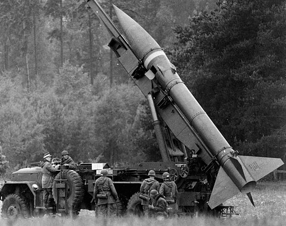 Eine MGR-1 Kurzstreckenrakete befindet sich in Feuerstellung auf einer Abschussvorrichtung, die auf einem LKW montiert ist. Vor dem LKW stehen Soldaten der Bundeswehr.