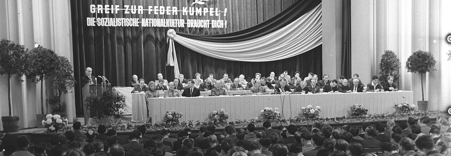 Blick in einen großen, voll besetzten und festlich geschmückten Konferenzsaal. Auf der Bühne befinden sich drei Reihen langer Tische, an denen jeweils 14 formal gekleidete Männer und einige Frauen sitzen. Am Rednerpult links spricht ein Mann. An der Wand hinter der Bühne steht der Aufruf 'Greif zur Feder Kumpel, die sozialistische Nationalkultur braucht Dich“.