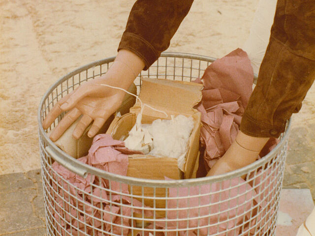 Eine Puppe beugt sich über einen metallenen Mülleimer, in dem Papier und eine Bombe liegen.