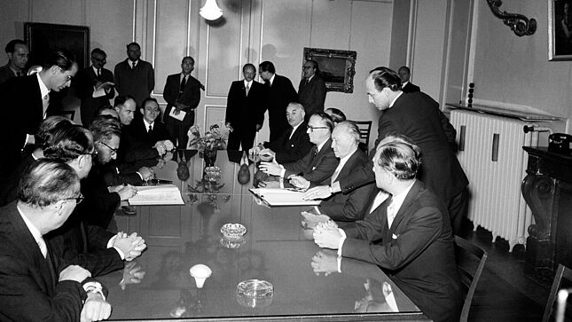 Konrad Adenauer und Mosche Sharet sitzen sich an einem Tisch gegenüber und haben jeweils eine geöffnete Vertragsmappe vor sich und einen Stift in der Hand. Weitere Männer in Anzügen sitzen mit am Tisch, einige stehen etwas abseits im Raum.