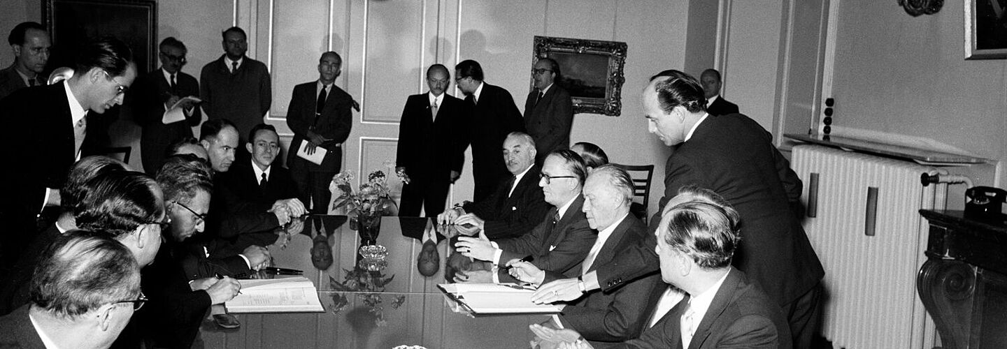 Unterzeichnung des Luxemburger Abkommens, 10. September 1952 Konrad Adenauer und Mosche Sharet sitzen sich an einem Tisch gegenüber und haben jeweils eine geöffnete Vertragsmappe vor sich und einen Stift in der Hand. Weitere Männer in Anzügen sitzen mit am Tisch, einige stehen etwas abseits im Raum.