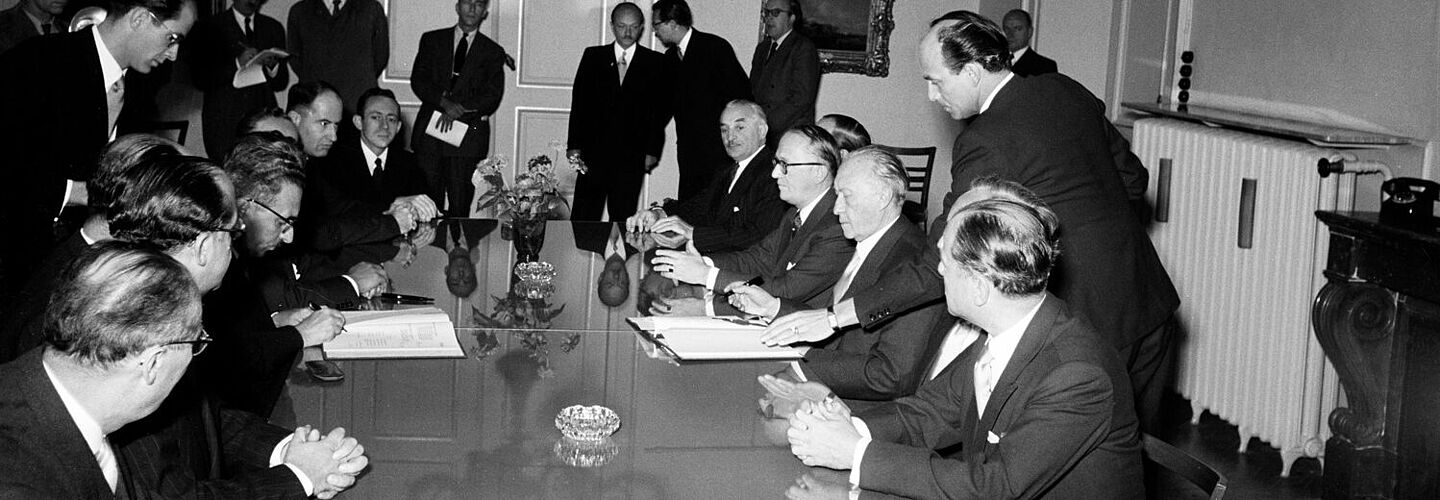 Konrad Adenauer und Mosche Sharet sitzen sich an einem Tisch gegenüber und haben jeweils eine geöffnete Vertragsmappe vor sich und einen Stift in der Hand. Weitere Männer in Anzügen sitzen mit am Tisch, einige stehen etwas abseits im Raum.