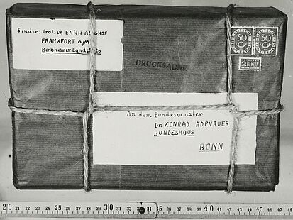 Aufgeklebtes Schwarz-Weiß-Foto eines mit Band verschnürten Pakets mit handgeschriebener Absender- und Empfängeradresse auf Etiketten, Briefmarken und „Drucksache“-Stempel; an einem darunter angelegten Zollstock ist das Maß ablesbar.