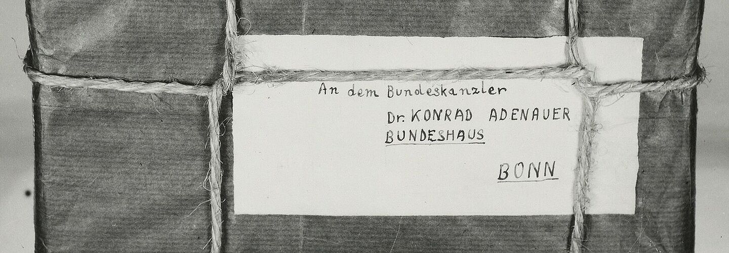 Aufgeklebtes Schwarz-Weiß-Foto eines mit Band verschnürten Pakets mit handgeschriebener Absender- und Empfängeradresse auf Etiketten, Briefmarken und „Drucksache“-Stempel; an einem darunter angelegten Zollstock ist das Maß ablesbar.