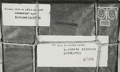 Aufgeklebtes Schwarz-Weiß-Foto eines mit Band verschnürten Pakets mit handgeschriebener Absender- und Empfängeradresse auf Etiketten, Briefmarken und „Drucksache“-Stempel; an einem darunter angelegten Zollstock ist das Maß ablesbar.