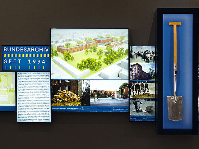 Beleuchtete Fotos und Texte sowie eine Vitrine mit einem Spaten sind an eine Wand montiert.