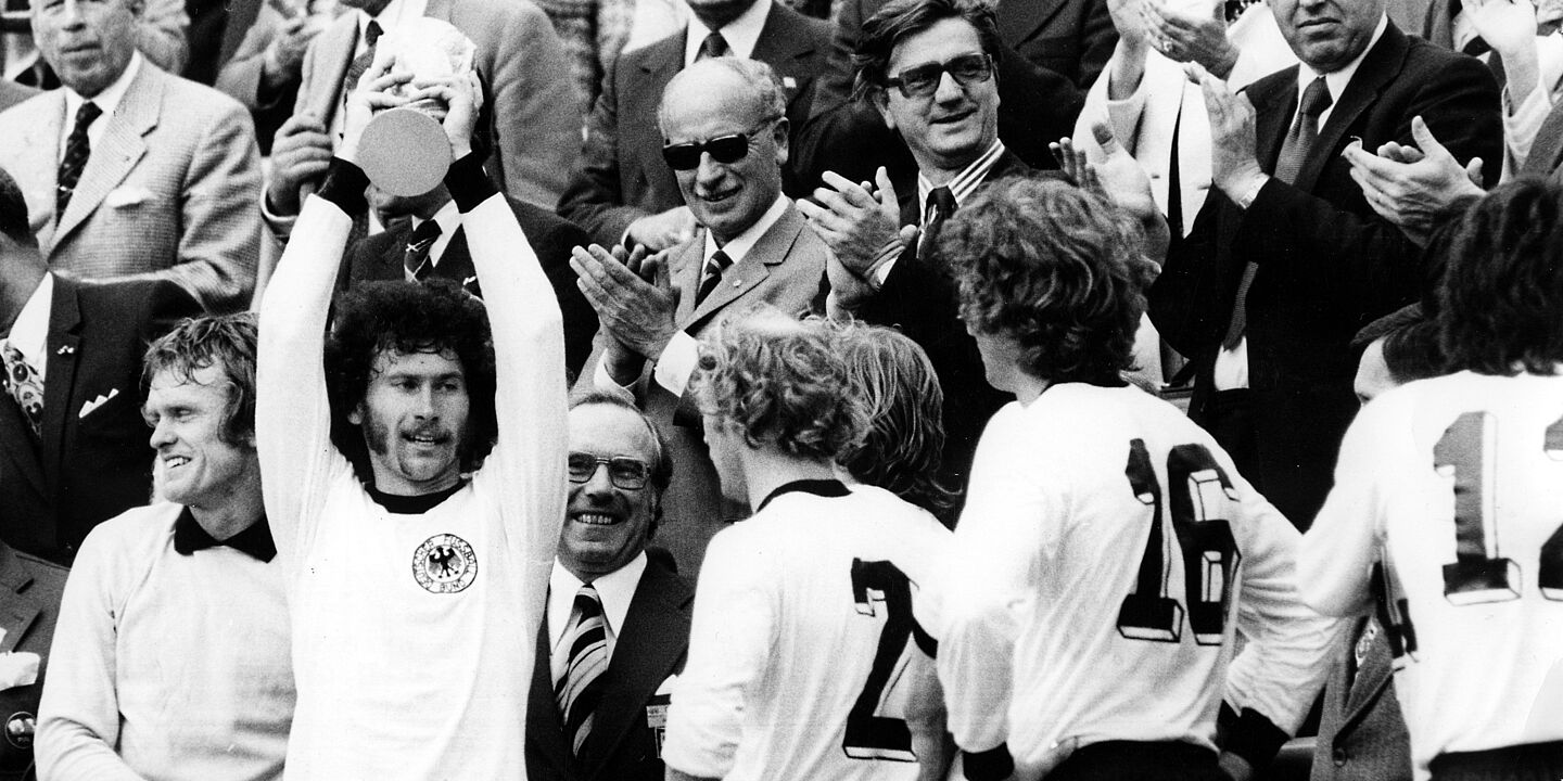 Paul Breitner hält auf der Tribüne mit beiden Händen den Pokal in die Luft. Weitere Spieler im BRD-Trikot stehen neben ihm. Mehrere Männer in Anzügen stehen dahinter, schauen zu und applaudieren., Quelle:
            BArch, Bild 183-N0716-317 / Mittelstädt, Rainer