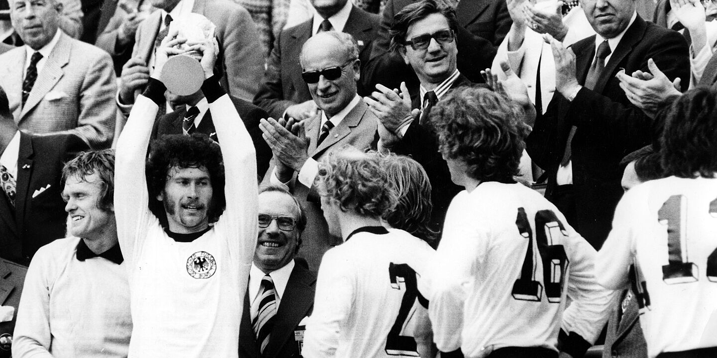 BRD-Spieler Paul Breitner hält den gewonnenen Welt-Cup hoch. Von links: Torwart Sepp Maier, Paul Breitner, Berti Vogts und Rainer Bonhof. Paul Breitner hält auf der Tribüne mit beiden Händen den Pokal in die Luft. Weitere Spieler im BRD-Trikot stehen neben ihm. Mehrere Männer in Anzügen stehen dahinter, schauen zu und applaudieren., Quelle:
BArch, Bild 183-N0716-317 / Mittelstädt, Rainer