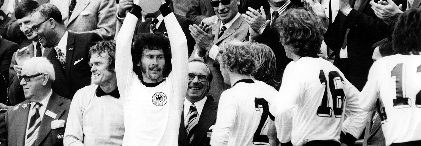 BRD-Spieler Paul Breitner hält den gewonnenen Welt-Cup hoch. Von links: Torwart Sepp Maier, Paul Breitner, Berti Vogts und Rainer Bonhof. Paul Breitner hält auf der Tribüne mit beiden Händen den Pokal in die Luft. Weitere Spieler im BRD-Trikot stehen neben ihm. Mehrere Männer in Anzügen stehen dahinter, schauen zu und applaudieren.