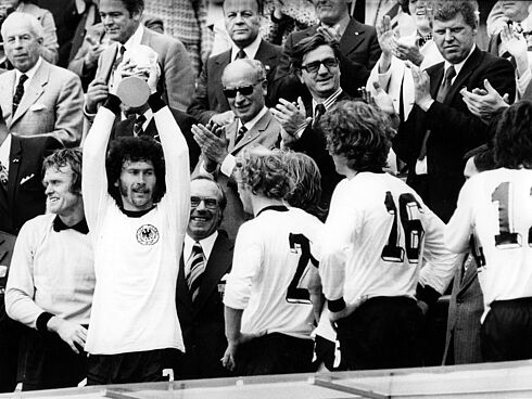 BRD-Spieler Paul Breitner hält den gewonnenen Welt-Cup hoch. Von links: Torwart Sepp Maier, Paul Breitner, Berti Vogts und Rainer Bonhof. Paul Breitner hält auf der Tribüne mit beiden Händen den Pokal in die Luft. Weitere Spieler im BRD-Trikot stehen neben ihm. Mehrere Männer in Anzügen stehen dahinter, schauen zu und applaudieren.