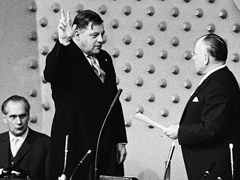 Das Schwarz-Weiß-Foto wurde am 14. November 1961 vom Fotografen Egon Steiner aufgenommen. Festgehalten wurde die Vereidigung von Franz Josef Strauß zum Verteidigungsminister. Strauß steht mit erhobener Hand und drei gestreckten Fingern gegenüber dem Präsidenten des Deutschen Bundestages, Eugen Gerstenmaier. Dieser hält ein Blatt Papier in der Hand. Im Hintergrund sitzt ein weiterer Herr im Anzug und verfolgt das Geschehen.