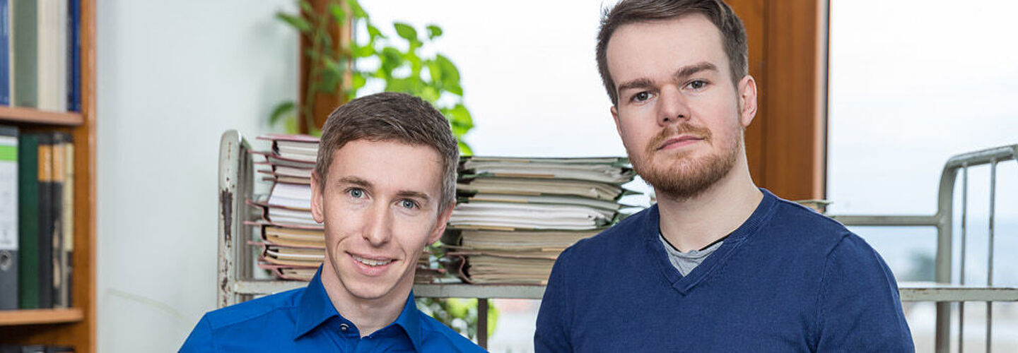 Zwei Männer halten Akten in den Händen und schauen lächeln in die Kamera.