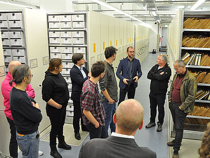 Männer und Frauen stehen in einem Magazinraum des Stasi-Unterlagen-Archivs und sprechen miteinander.