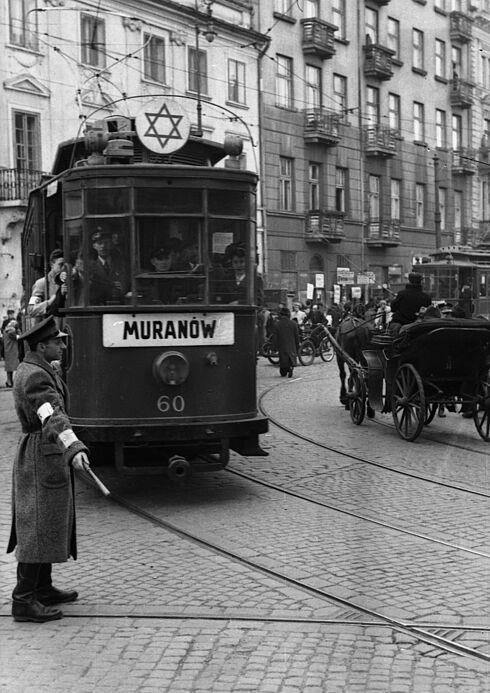 Diese Schwarz-Weiß-Fotografie zeigt eine im Warschauer Ghetto fahrende Straßenbahn der Linie 60 in Richtung Muranow. In der Straßenbahn befinden sich neben dem Fahrzeugführer auch Fahrgäste. Des Weiteren ist sie mit einem Judenstern gekennzeichnet. Mitten auf der Kreuzung steht ein Mitglied des jüdischen Ordnungsdienstes, welches offenbar den Verkehr regelt. Auf der anderen Straßenseite fährt eine Kutsche.