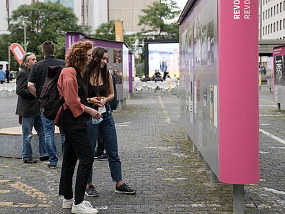 Zwei junge Frauen stehen auf den Innenhof der ehemaligen Stasi-Zentrale und sehen sich eine große Schautafel der Open-Air-Ausstellung "Revolution und Mauerfall" an