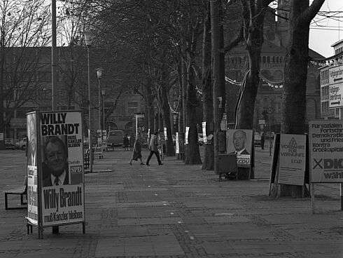 Schwarz-Weiß-Foto mit an Bäumen und Laternen aufgestellten Wahlkampfplakaten, u. a. mit dem Gesicht von Willy Brandt