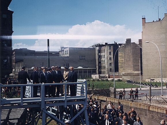 John F. Kennedy steht in einer kleinen Gruppe auf einer Plattform am Checkpoint Charlie unmittelbar an der Berliner Mauer mit Blick auf den Ostteil der Stadt. Neben ihm steht Bundeskanzler Konrad Adenauer. Unten befindet sich eine Menschenansammlung, darunter Journalisten. 