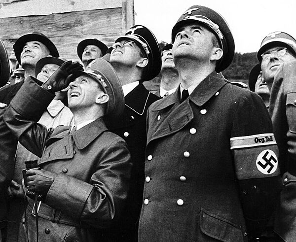 Das schwarzweiße Gruppenfoto zeigt mehrere Männer in Uniformen, die nach oben schauen. 