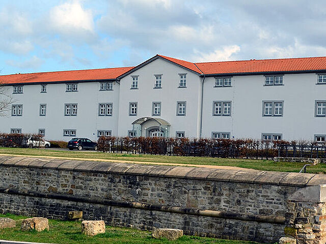 Das Bundesarchiv – Stasi-Unterlagen-Archiv Erfurt (Nordansicht): Im Vordergrund eine leere Straße, dahinter ein grüner Parkplatz und ein langgezogenes weißes Gebäude nmit 3 Stöcken und einem roten flachen Satteldach. Der ist Blau mit weißen Wölkchen.