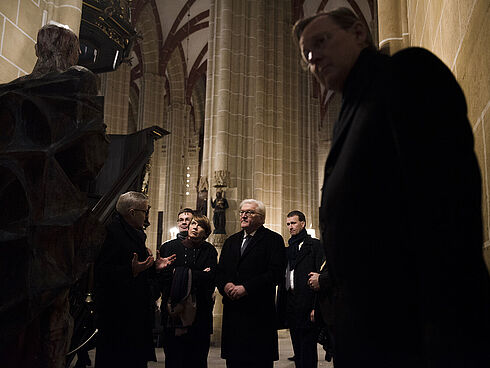 Mehrere Personen, darunter Frank-Walter Steinmeier, Elke Büdenbender und Bodo Ramelow, stehen in einer Kirche und hören einem Mann zu.