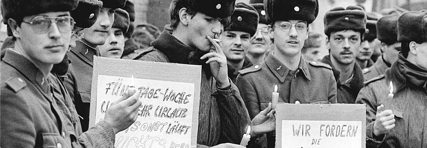 Junge Soldaten der NVA in Uniform protestieren 1990 in Cottbus für bessere Dienstbedingungen. Die Soldaten tragen Schilder mit den Aufschriften „Fünf Tage-Woche und mehr Urlaub sonst läuft nichts mehr“ und „Wir fodern die Verkürzung des Wehrdienstes auf 12 Monate! *rückwirkend“. Einige Soldaten haben Kerzen angezündet.
