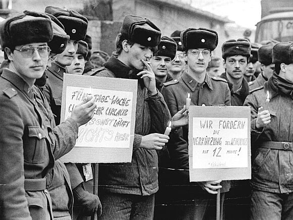 Junge Soldaten der NVA in Uniform protestieren 1990 in Cottbus für bessere Dienstbedingungen. Die Soldaten tragen Schilder mit den Aufschriften „Fünf Tage-Woche und mehr Urlaub sonst läuft nichts mehr“ und „Wir fodern die Verkürzung des Wehrdienstes auf 12 Monate! *rückwirkend“. Einige Soldaten haben Kerzen angezündet.