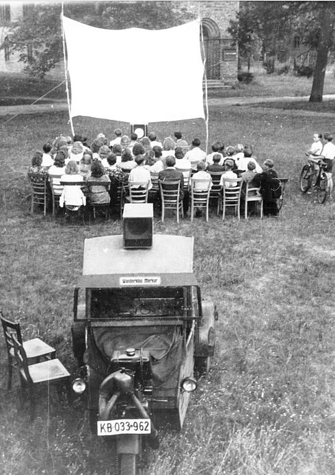 Etwa 50 Personen sitzen auf Stühlen auf einer Wiese und schauen auf eine provisorisch befestigte Leinwand. Lautsprecher und Projektionstechnik befinden sich auf einem dreirädrigen Gefährt, das hinter den Stuhlreihen steht.