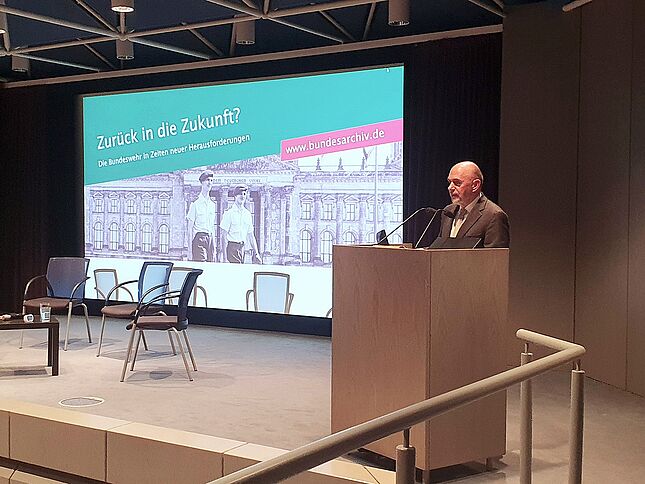 Michael Steidel bei einer Veranstaltung zum Thema Bundeswehr Ein Mann im Anzug steht an einem Pult auf einer Bühne und spricht ins Mikrofon. Im Hintergrund wird ein Bild projiziert mit den Worten „Zurück in die Zukunft? Die Bundeswehr in Zeiten neuer Herausforderungen“.