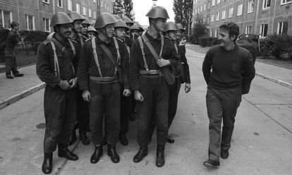 In Reihe marschierende Soldaten in NVA-Uniform blicken auf einen Kameraden, der bereits Bundeswehrausrüstung trägt.