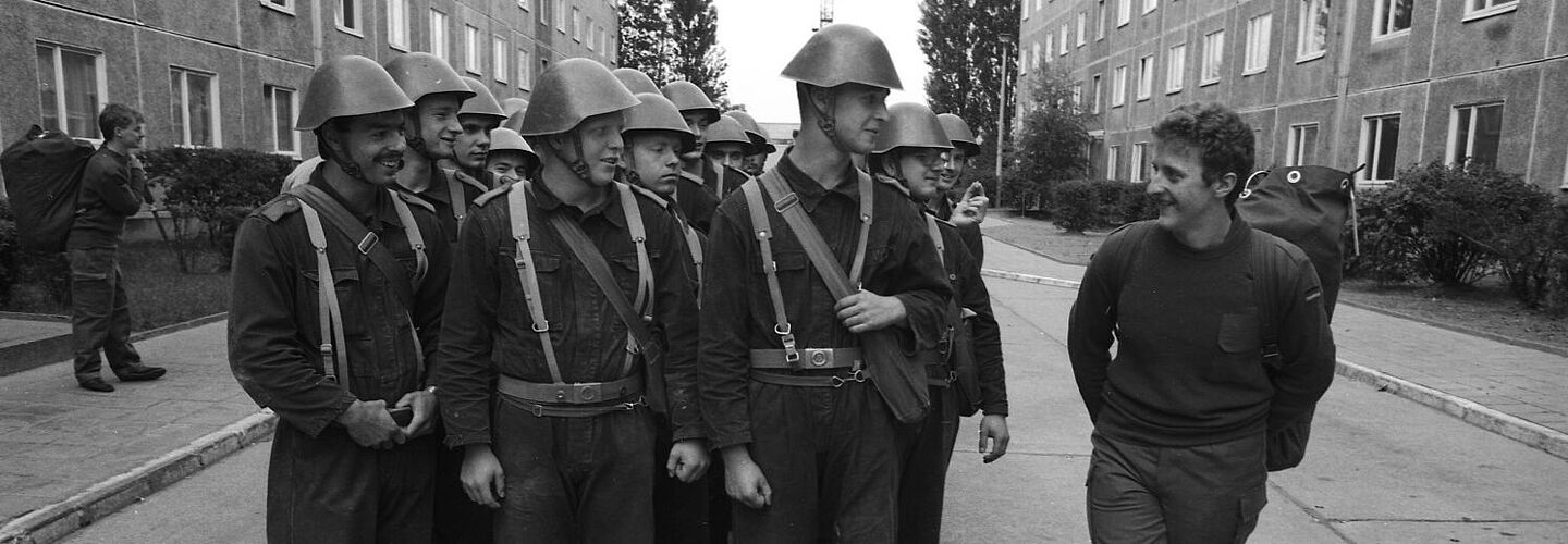 In Reihe marschierende Soldaten in NVA-Uniform blicken auf einen Kameraden, der bereits Bundeswehrausrüstung trägt.