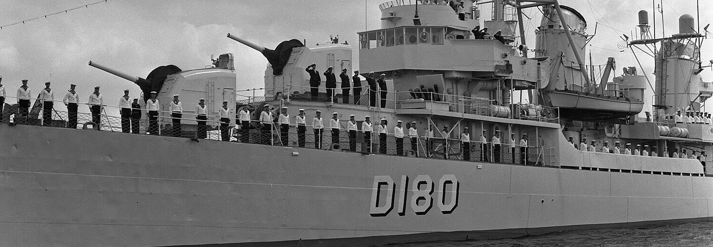 Kriegsschiff, auf dem mehrere Matrosen aufgereiht stehen.