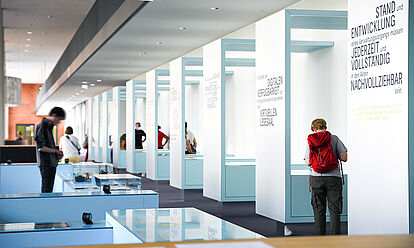 Besucherinnen und Besucher betrachten Ausstellungstische und -vitrinen in der Ausstellung „Geschichte in Geschichten“.