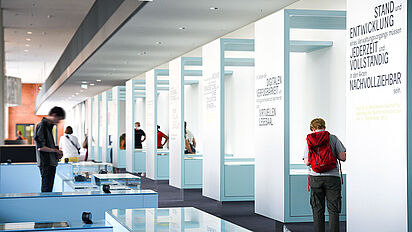 Besucherinnen und Besucher betrachten Ausstellungstische und -vitrinen in der Ausstellung „Geschichte in Geschichten“., Quelle:
                                                Bundesarchiv / Körner