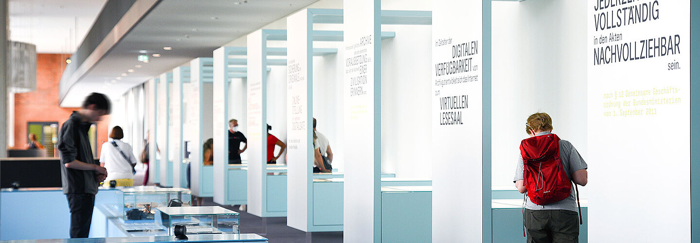 Besucherinnen und Besucher betrachten Ausstellungstische und -vitrinen in der Ausstellung „Geschichte in Geschichten“.