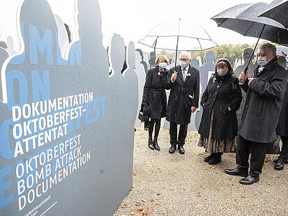 Mehrere Personen mit Mund-Nasen-Schutz und Schirmen stehen vor der Dokumentationsstätte zum Oktoberfestattentat.