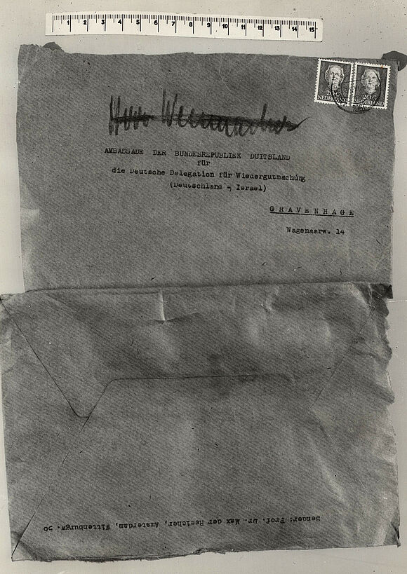 Zwei untereinander aufgeklebte Paketpapierseiten, die obere maschinell beschriftet mit der Adresse der deutschen Botschaft in Den Haag sowie einem händisch geschriebenen, durchgestrichenen Namen und zwei niederländischen Briefmarken; die untere mit Amsterdamer Absenderangaben.