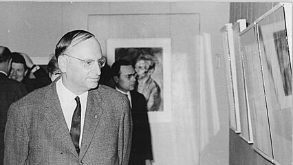 Kurt Hager schaut sich in einer Berliner Ausstellung ein Exponat an, das an der Wand hängt. Er trägt einen Anzug und eine Krawatte.