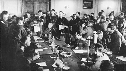 Wilhelm Keitel sitzt während der Unterzeichnung der deutschen Kapitulationserklärung an einem Tisch. Er ist umringt von Personen, darunter Georgi Schukow.